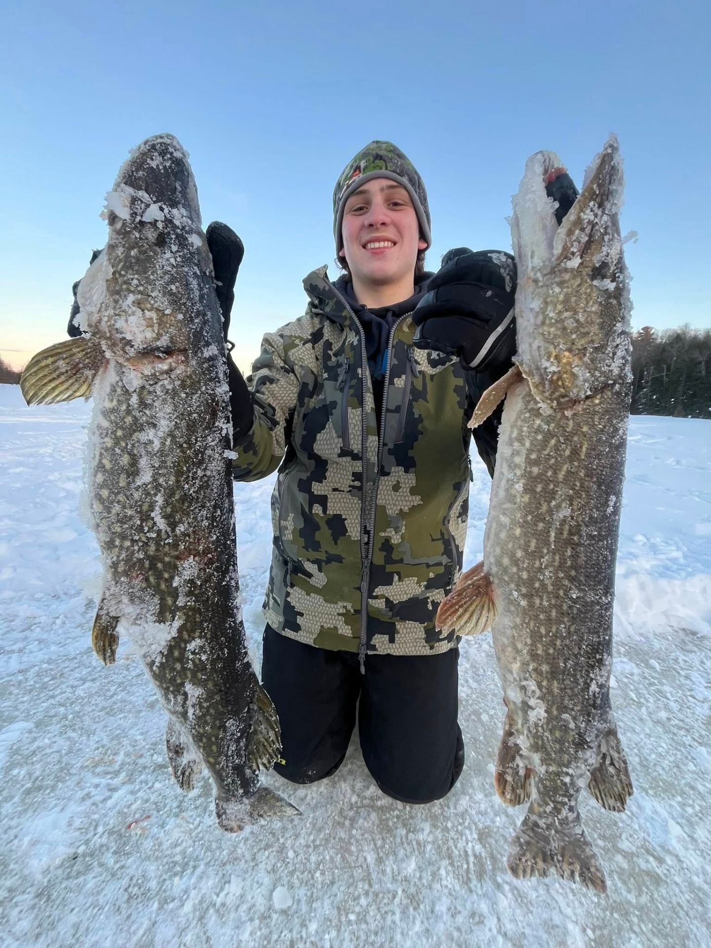 Angler holding up two large northern pike caught ice fishing on a frozen lake at sunrise